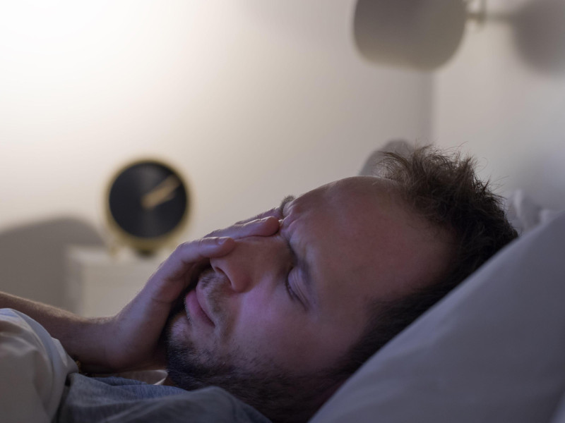 Concepto de insomnio y pesadillas. Un hombre no puede dormir después de una fiesta, se acuesta en la cama por la noche y tiene el despertador sobre la mesilla de noche. Trastornos del ciclo sueño/vigilia