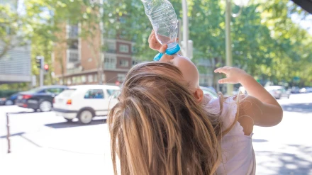 Espalda de un niño rubio caucásico de cuatro años con camisa blanca bebiendo de una botella de agua de plástico vacía, en una calle de Madrid
